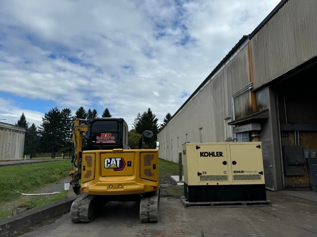 CAT excavator and Kohler generator near industrial building under a cloudy sky.