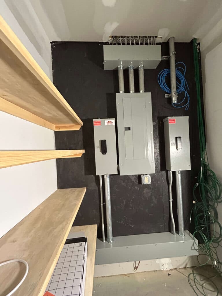 Electrical panel and wiring setup with wooden shelves in a residential utility room.