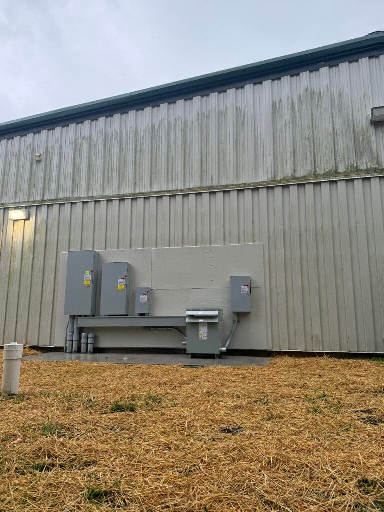Electrical equipment mounted on a gray wall beside a building with straw-covered ground.