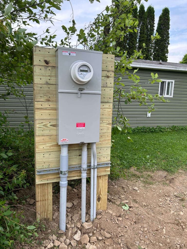 Electrical meter mounted on wooden post with conduit pipes in grassy area near a house.