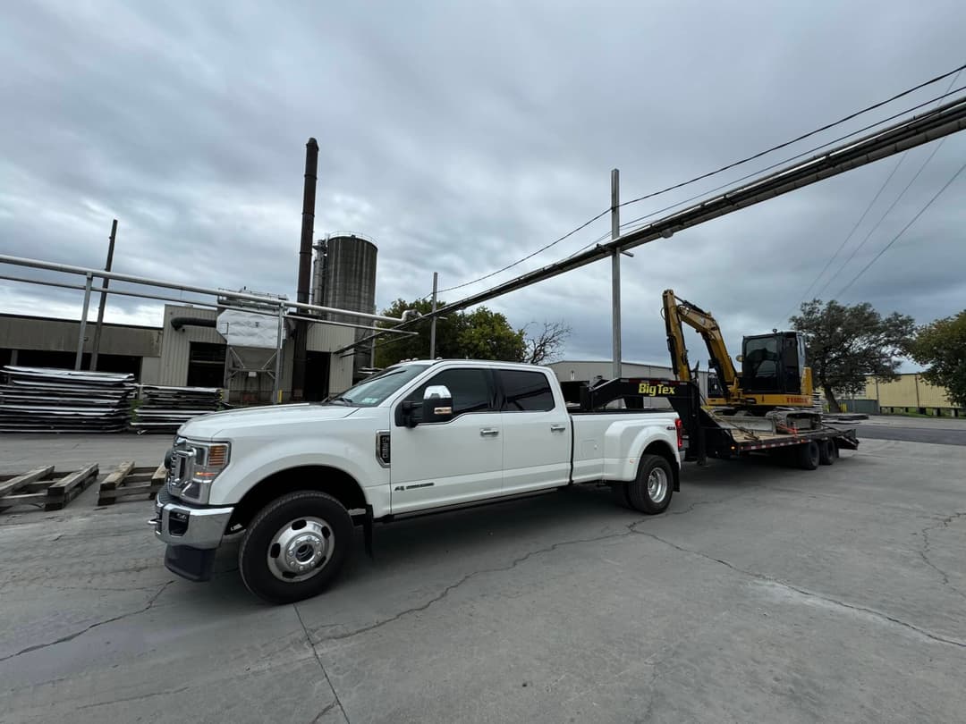 White pickup truck with a trailer carrying construction equipment in an industrial setting.