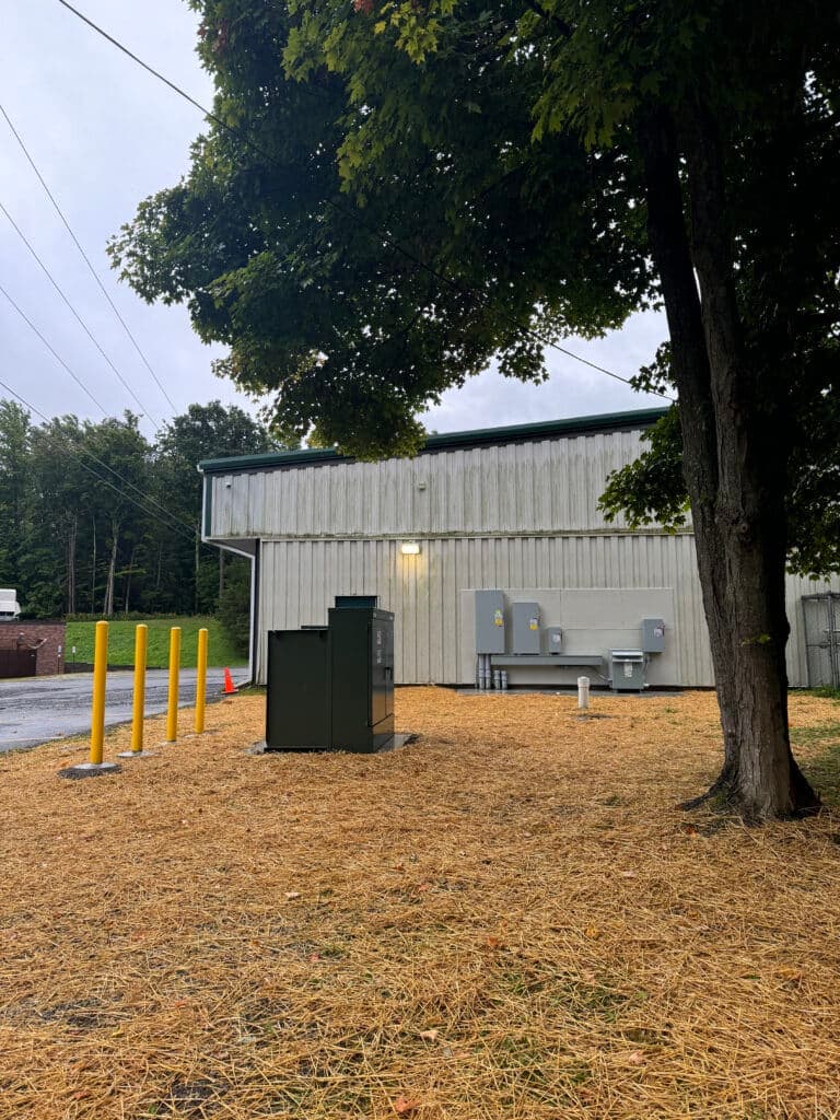 Utility building with utility equipment, surrounded by straw and a tree, on a cloudy day.