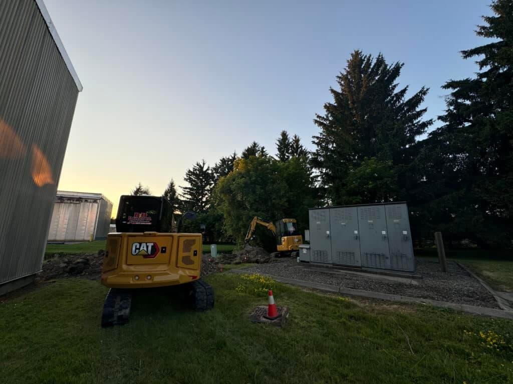 Excavation equipment near utility boxes and trees at a construction site during sunset.